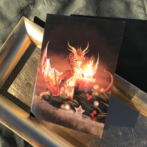A little read dragon uses his flame breath to light a festive candle on the friont of this Holiday Card